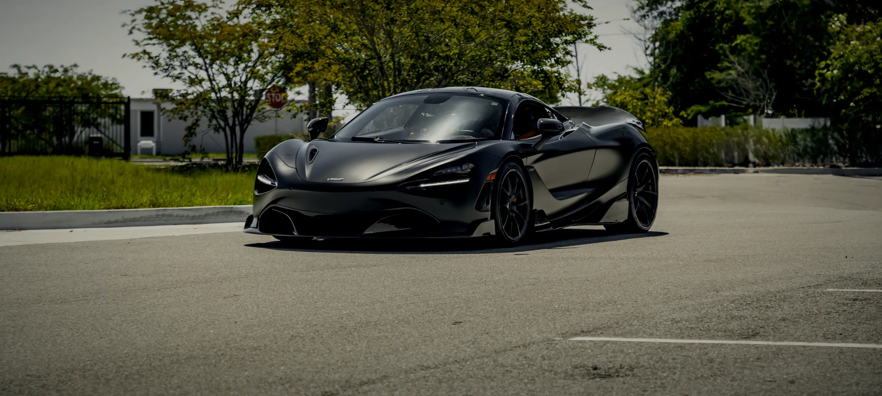 A dark McLaren parked in an open lot with trees behind it