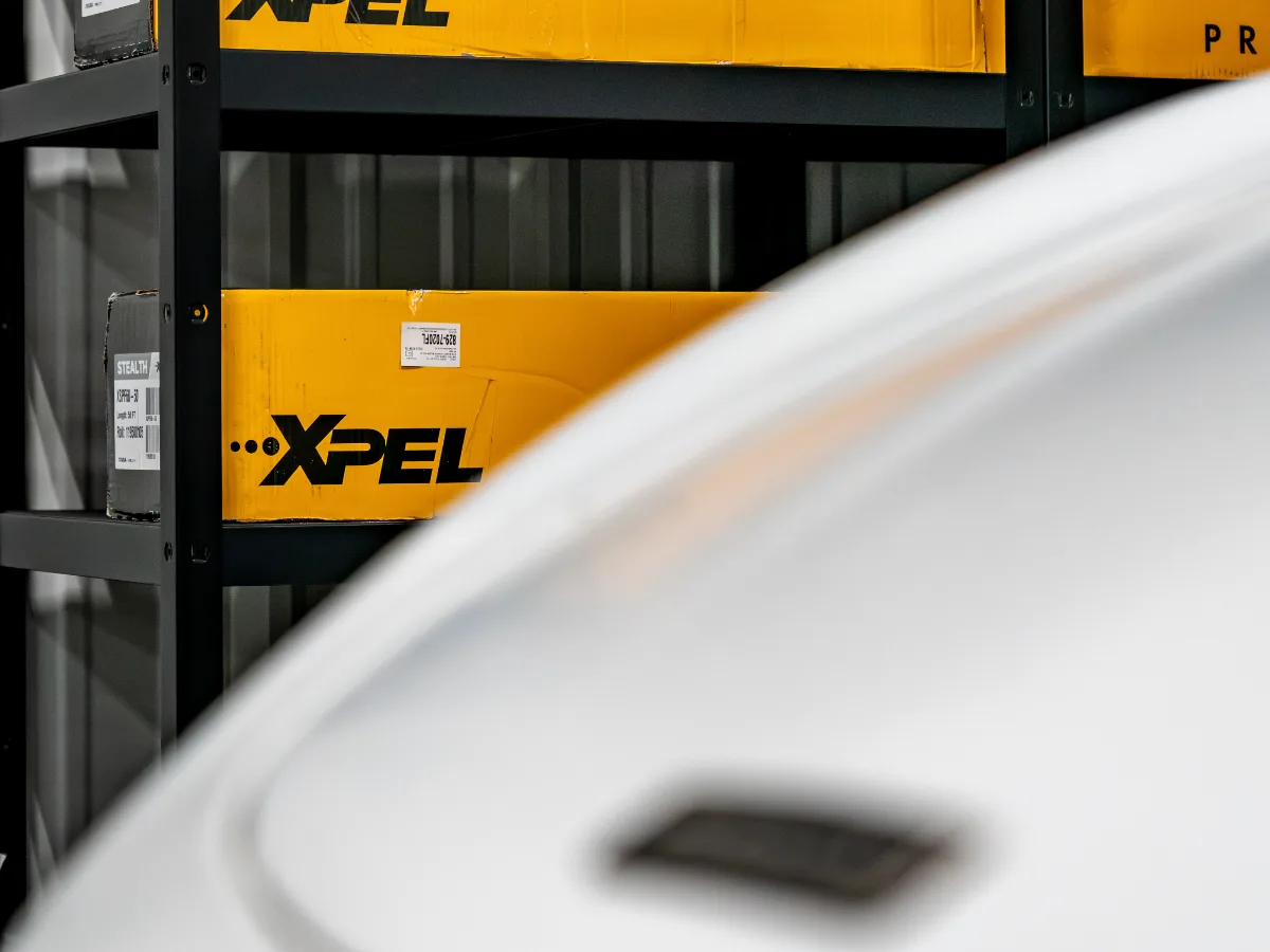 Boxed XPEL film stock shelved beside a car in the Detail Geex studio